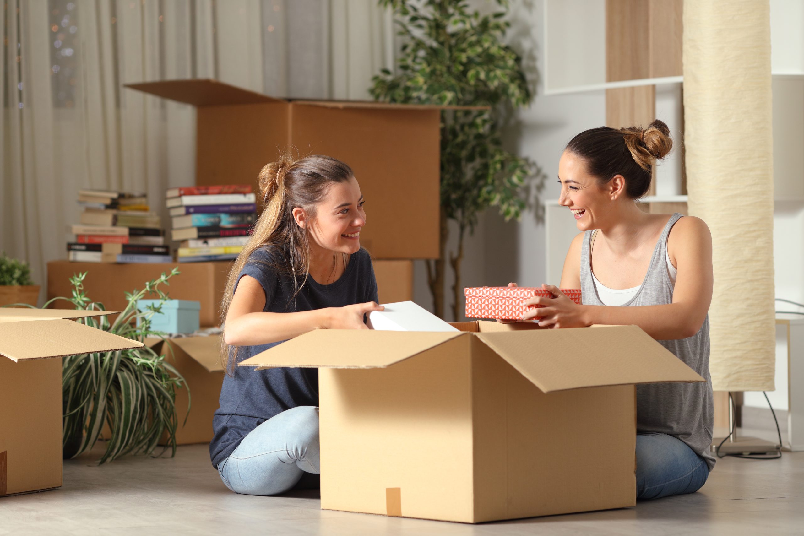 Zwei Frauen packen gemeinsam Umzugskartons in einer freundlichen Umgebung einer Kinder- & Jugendhilfeeinrichtung, umgeben von Büchern und Pflanzen, und lächeln glücklich miteinander.