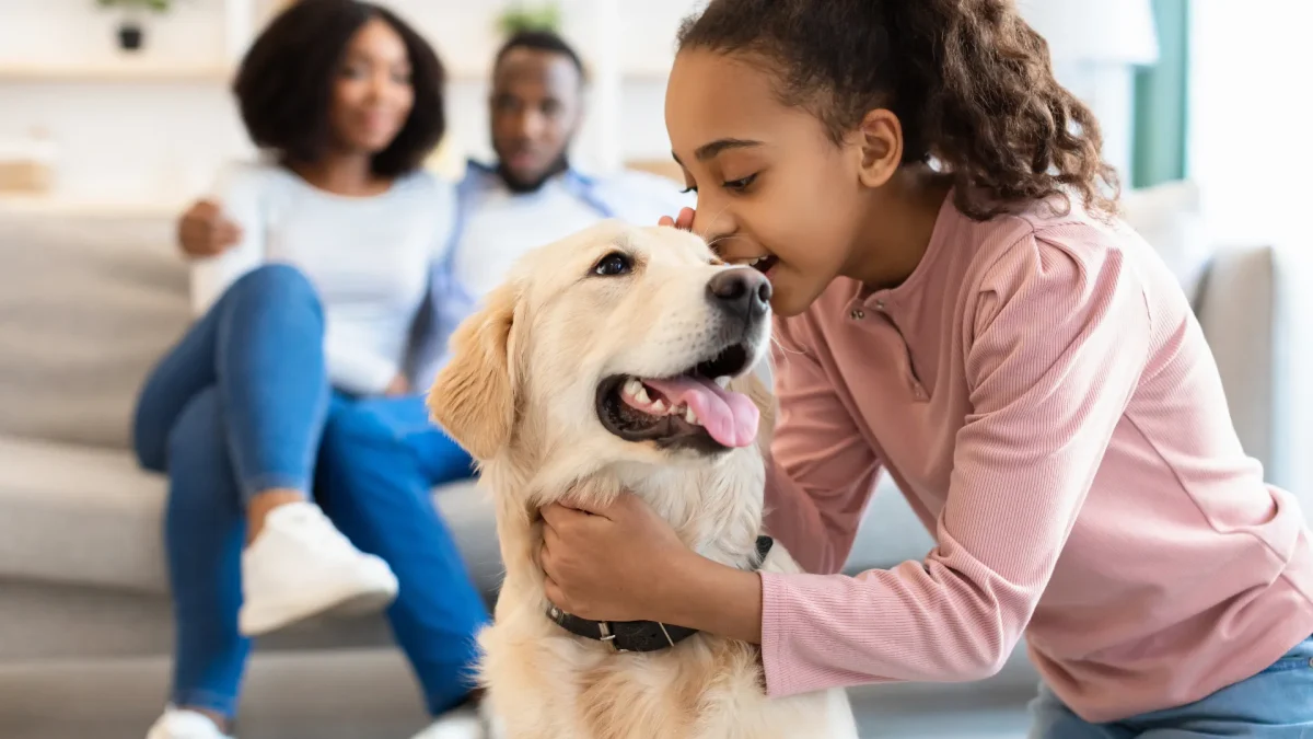 Mädchen streichelt Hund in einer liebevollen Umgebung, Eltern im Hintergrund. Glücklicher Moment in einer Kinder- & Jugendhilfeeinrichtung, förderndes Umfeld und tiergestützte Unterstützung.