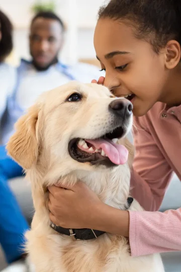 Mädchen streichelt Hund in einer liebevollen Umgebung, Eltern im Hintergrund. Glücklicher Moment in einer Kinder- & Jugendhilfeeinrichtung, förderndes Umfeld und tiergestützte Unterstützung.