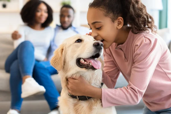 Mädchen streichelt Hund in einer liebevollen Umgebung, Eltern im Hintergrund. Glücklicher Moment in einer Kinder- & Jugendhilfeeinrichtung, förderndes Umfeld und tiergestützte Unterstützung.