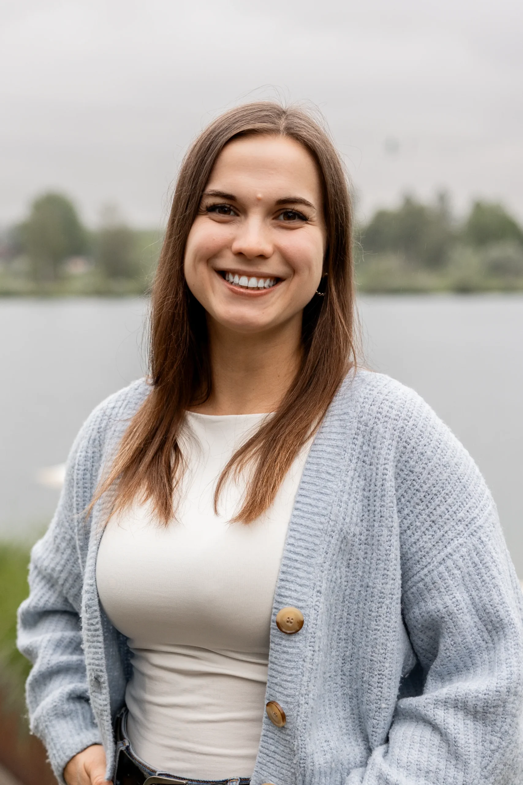 Eine junge Frau mit braunen Haaren lächelt und steht im Freien vor einem See, trägt einen hellen Pullover und ein weißes Oberteil.